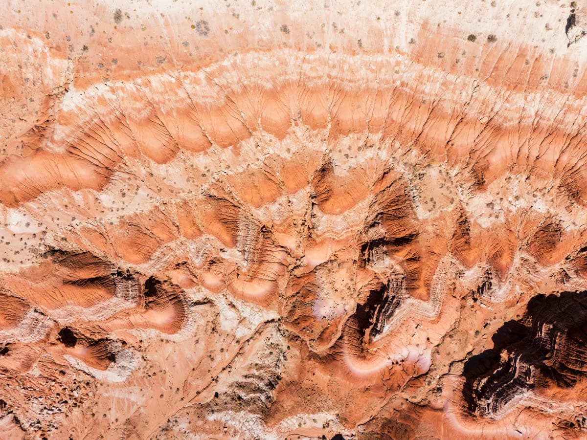 An overhead drone shot captures a mesmerizing pattern of striped badlands in the Utah desert. Swirls of red, pink, and white rock form wave-like ridges and valleys, revealing the complex erosion and natural texture of the landscape.