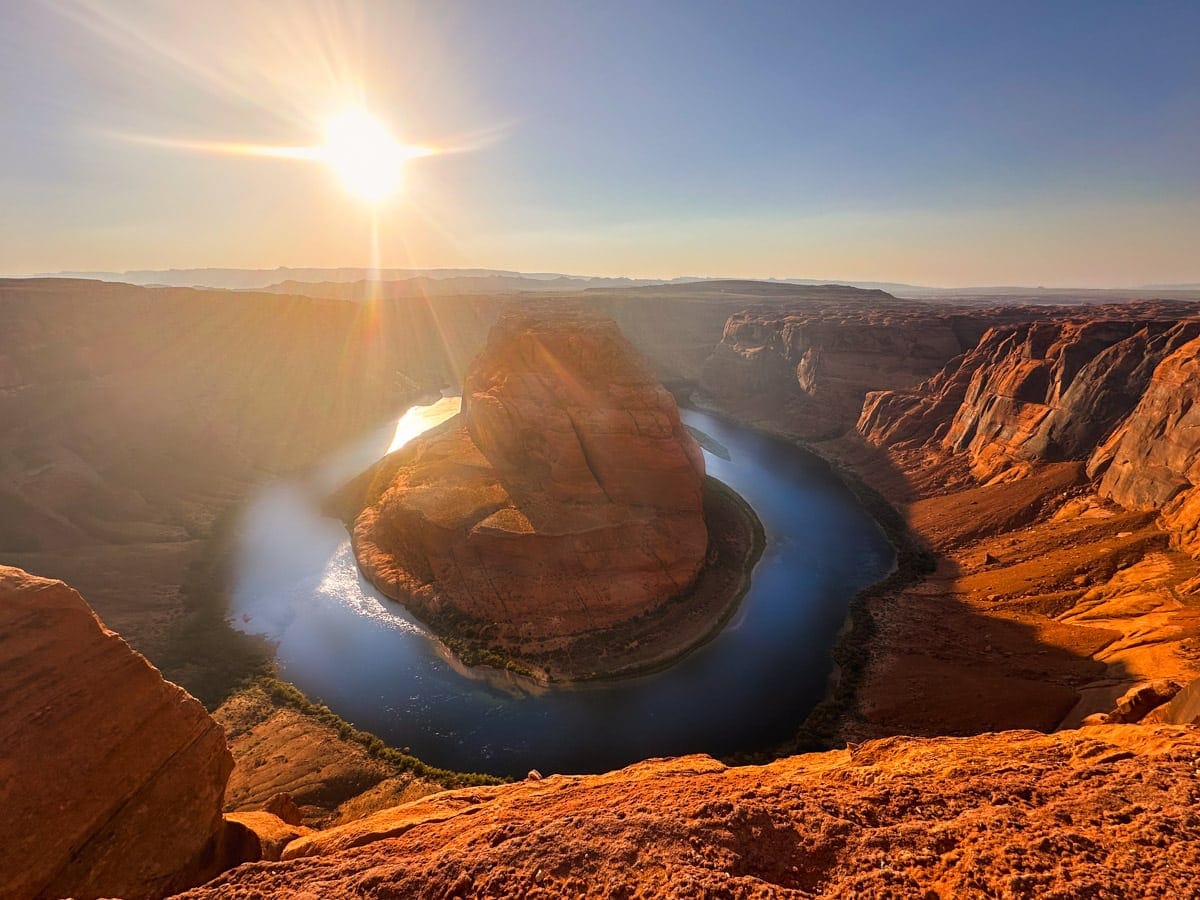Golden sunlight spills across the sandstone cliffs of Horseshoe Bend, where the Colorado River makes a dramatic U-shaped curve below. The water glows with soft blue and silver tones as the orange canyon walls reflect the warm rays of the setting sun.