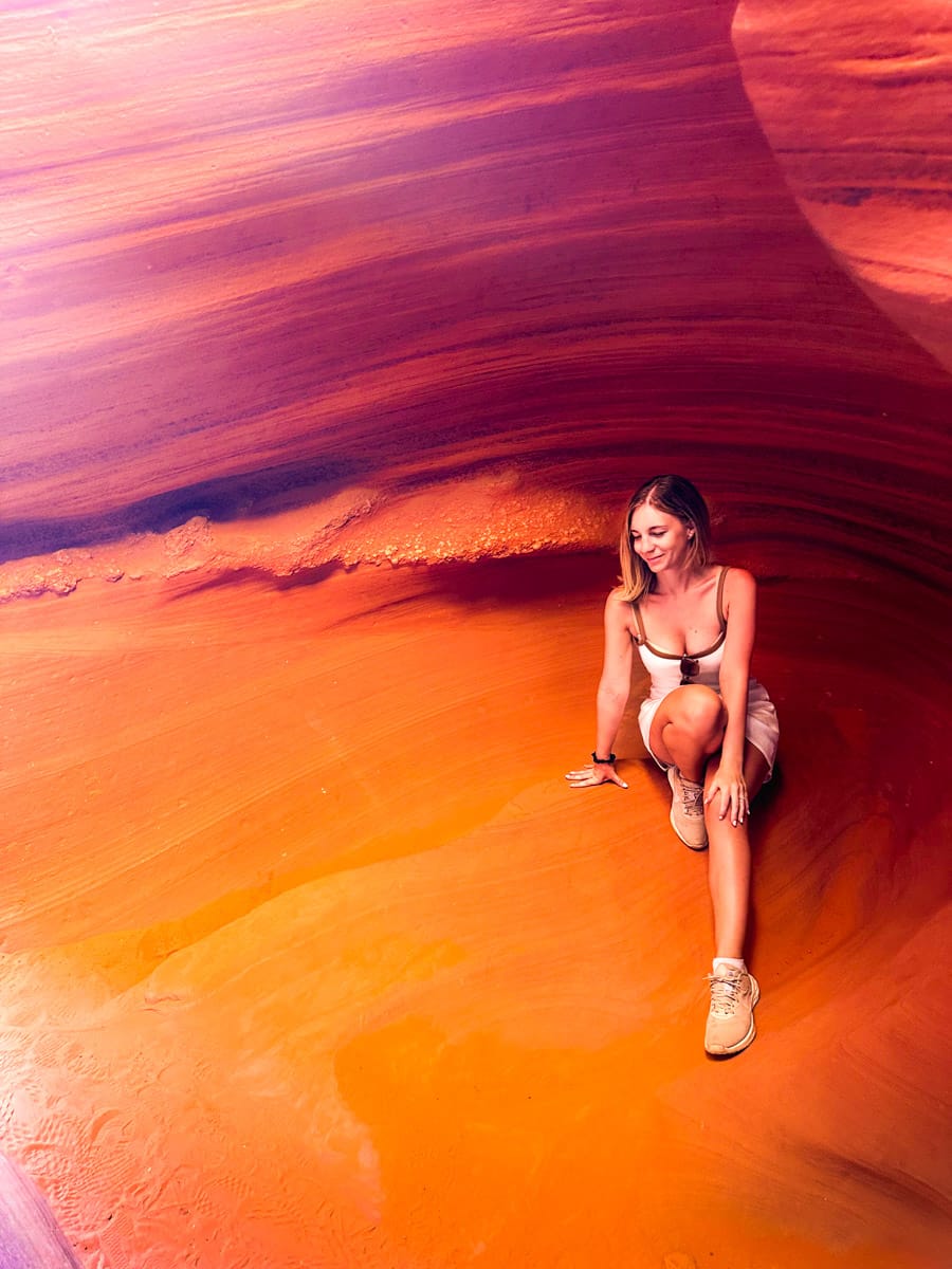 A woman sits gracefully inside a smooth, glowing chamber of Antelope Canyon, surrounded by rich shades of orange and violet sandstone. Her light dress contrasts with the warm rock, and soft natural light enhances the surreal and artistic beauty of the slot canyon.