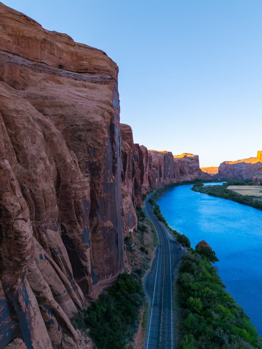 A scenic drive hugs the edge of the Colorado River in Utah, surrounded by towering red cliffs that glow with sunset light. The blue river contrasts sharply with the rich desert hues, making this a dream route for road trippers and nature lovers.