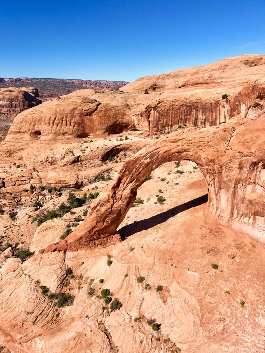 A drone view of the magnificent Corona Arch near Moab, Utah, rising from the red sandstone landscape in a sweeping curve. The arch casts a dramatic shadow, showing off its size and natural elegance under a bright, cloudless sky.