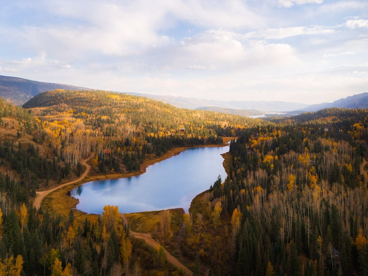 A peaceful mountain lake surrounded by a forest of aspens in full fall color, reflecting golden hues and a soft sky. Winding dirt roads and cabins are nestled among the trees, perfect for a cozy getaway.