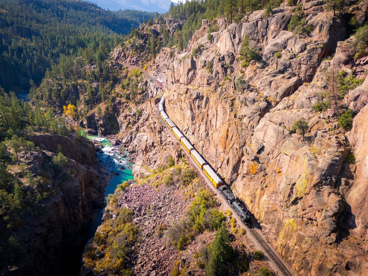 The historic Durango & Silverton Narrow Gauge Railroad winds through a rugged canyon, passing over the turquoise Animas River. Surrounded by steep cliffs and dense pine forest, the train looks straight out of a storybook.