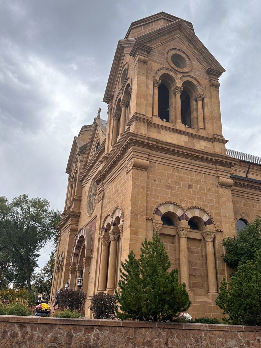 An upward view of the historic Cathedral Basilica of St. Francis of Assisi in Santa Fe, New Mexico, with its Romanesque architecture, carved columns, and soft sandstone tones standing out under a gray, moody sky.