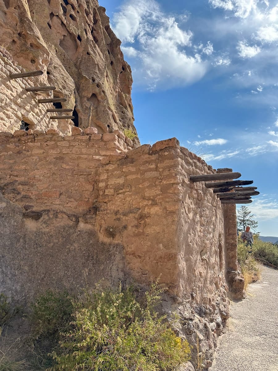 Ancient Puebloan ruins at Bandelier National Monument, New Mexico – Adobe cliff dwellings built into the rock face, with a clear blue sky above and a pathway leading toward the ruins.