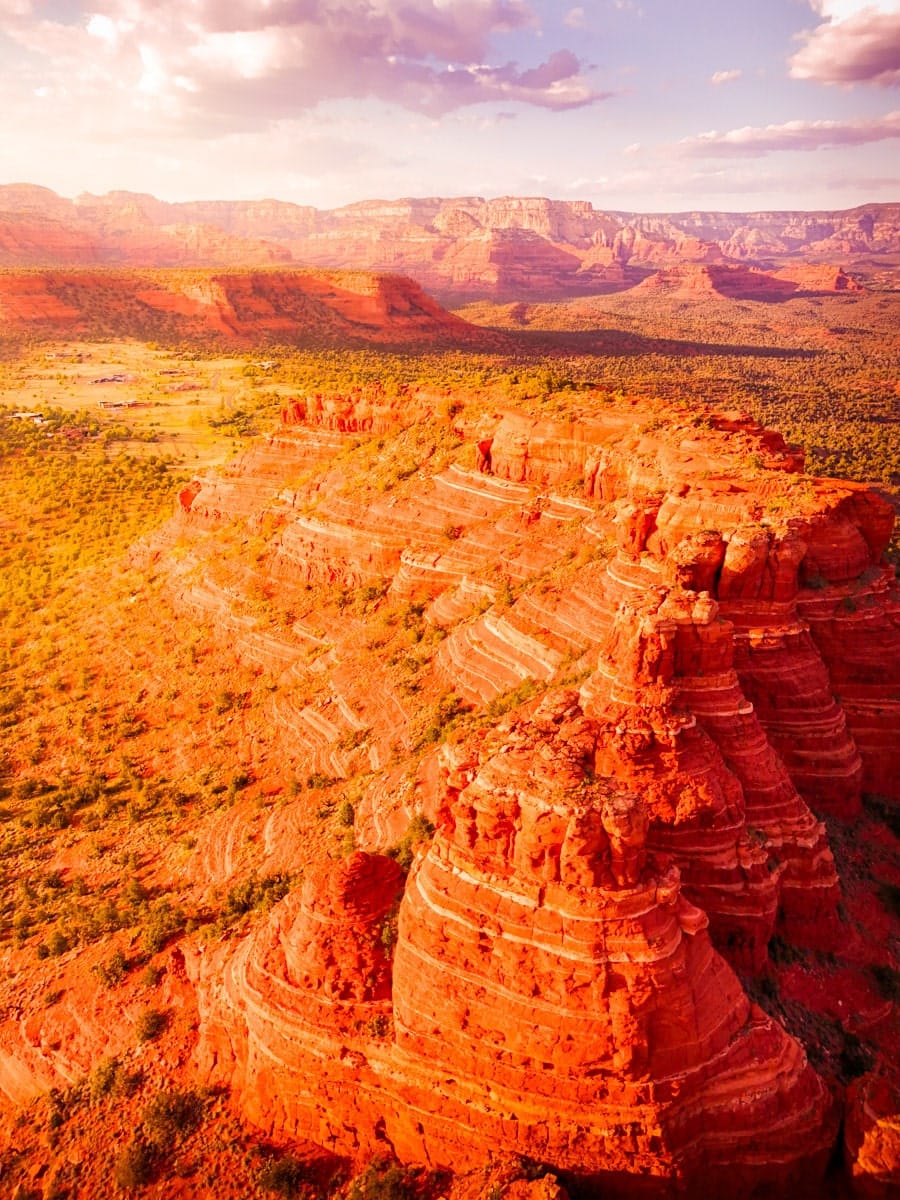 A vibrant aerial view of Sedona’s famous red rock formations at sunset, with dramatic layers of orange, red, and pink stone rising from a lush valley. The warm golden hour light bathes the entire scene in a fiery glow, making the textured cliffs and mesas even more jaw-dropping.