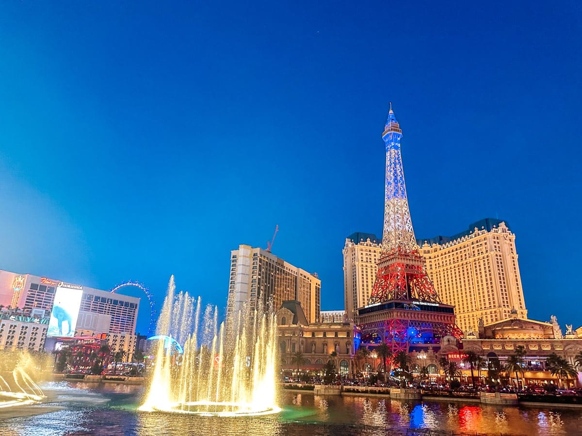 The Bellagio fountains burst into a glowing water show on the Las Vegas Strip, with the illuminated replica of the Eiffel Tower and Paris Las Vegas Hotel shining in the background. The bright lights and deep blue sky add extra dazzle to the iconic nighttime cityscape.
