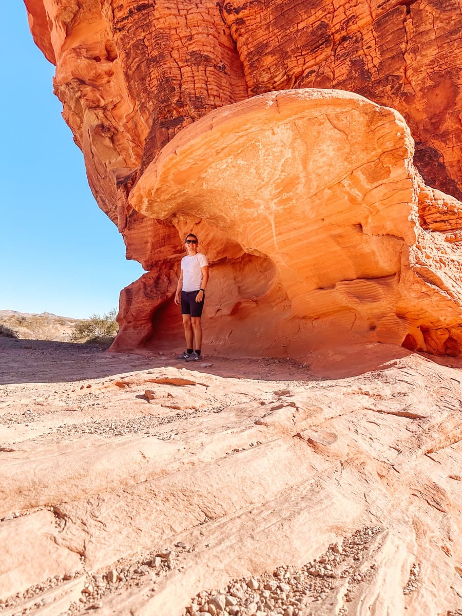 A man in sunglasses stands under a massive, curving red rock formation in Valley of Fire State Park, Nevada. The sandstone arch glows with deep orange tones as it towers over the desert floor, creating a dramatic contrast with the bright blue sky.