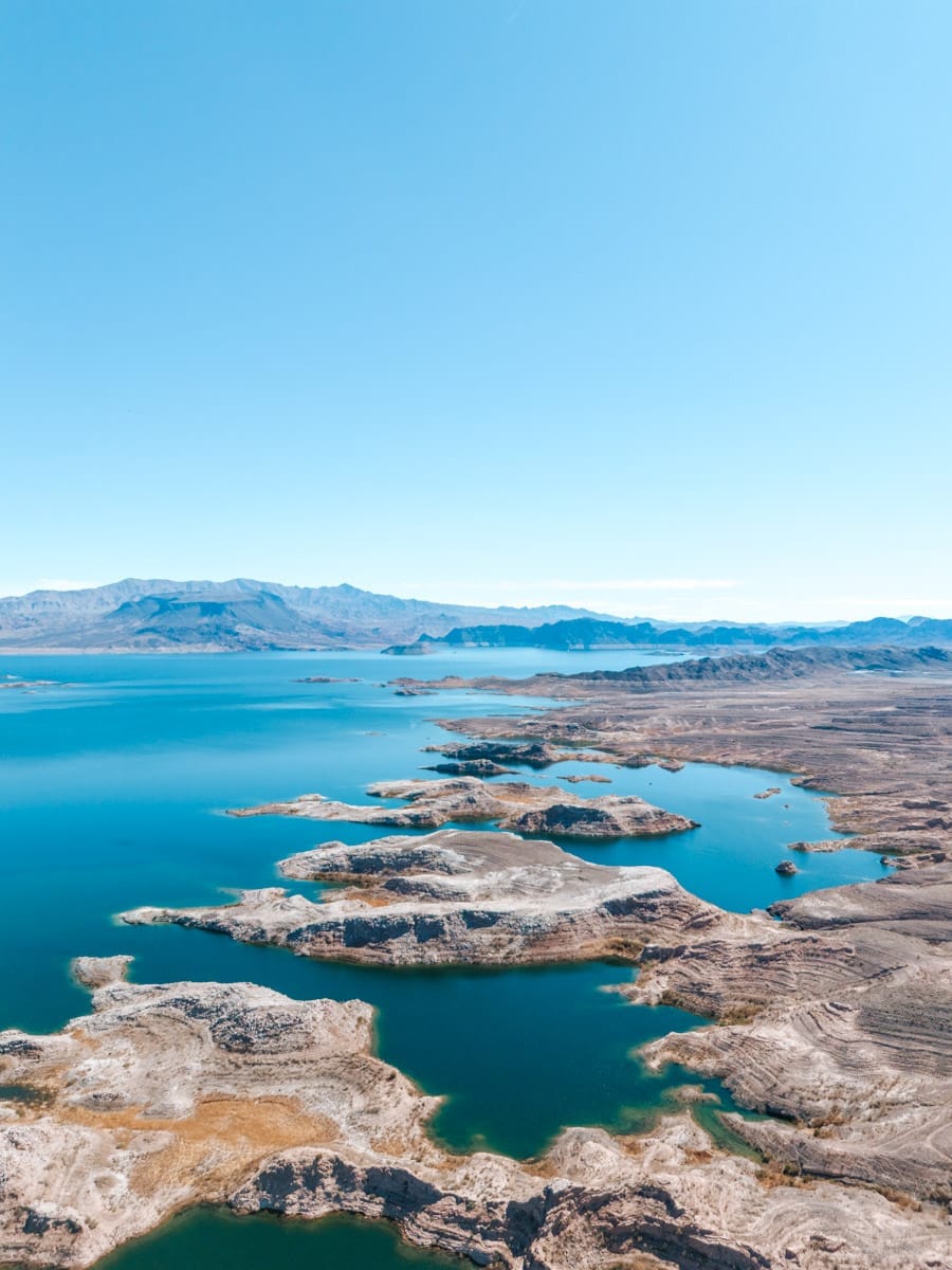 A stunning aerial shot of Lake Mead near the Nevada-Arizona border, featuring blue waters winding through dry rocky terrain. The contrast between the turquoise lake and the surrounding desert cliffs creates a striking natural pattern under a clear sky.