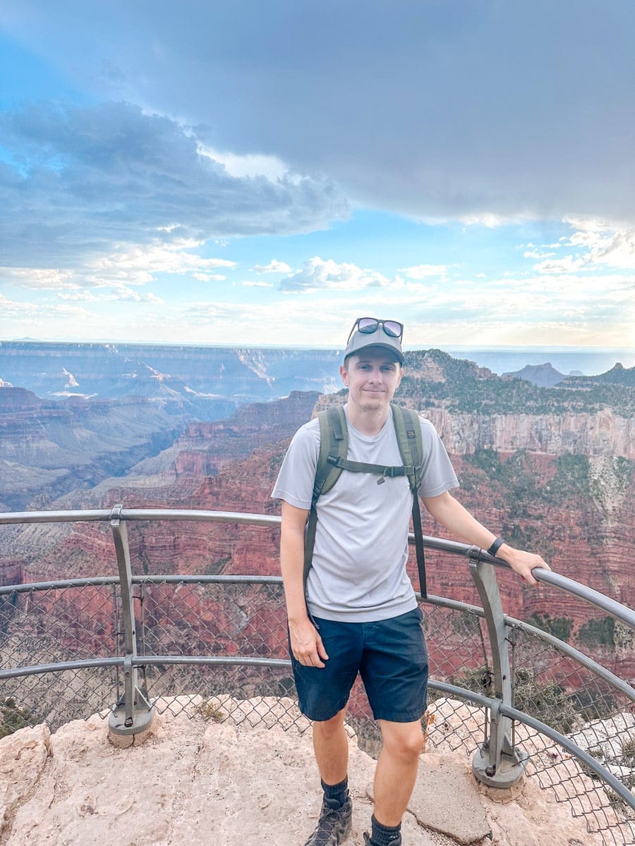 the author eans against a metal railing at a viewpoint on the North Rim of the Grand Canyon, surrounded by layers of red and orange cliffs stretching deep into the distance. Puffy clouds float above as he smiles, soaking in the epic views.