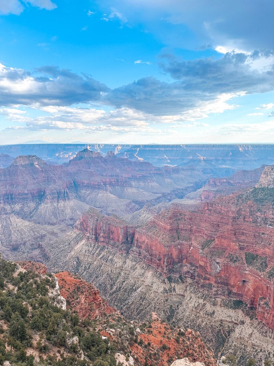 A vast, breathtaking view from the North Rim of the Grand Canyon shows dramatic red rock cliffs and deep canyons fading into the horizon. The scene is lit by soft daylight under a sky full of cotton-like clouds and mountain silhouettes in the distance.