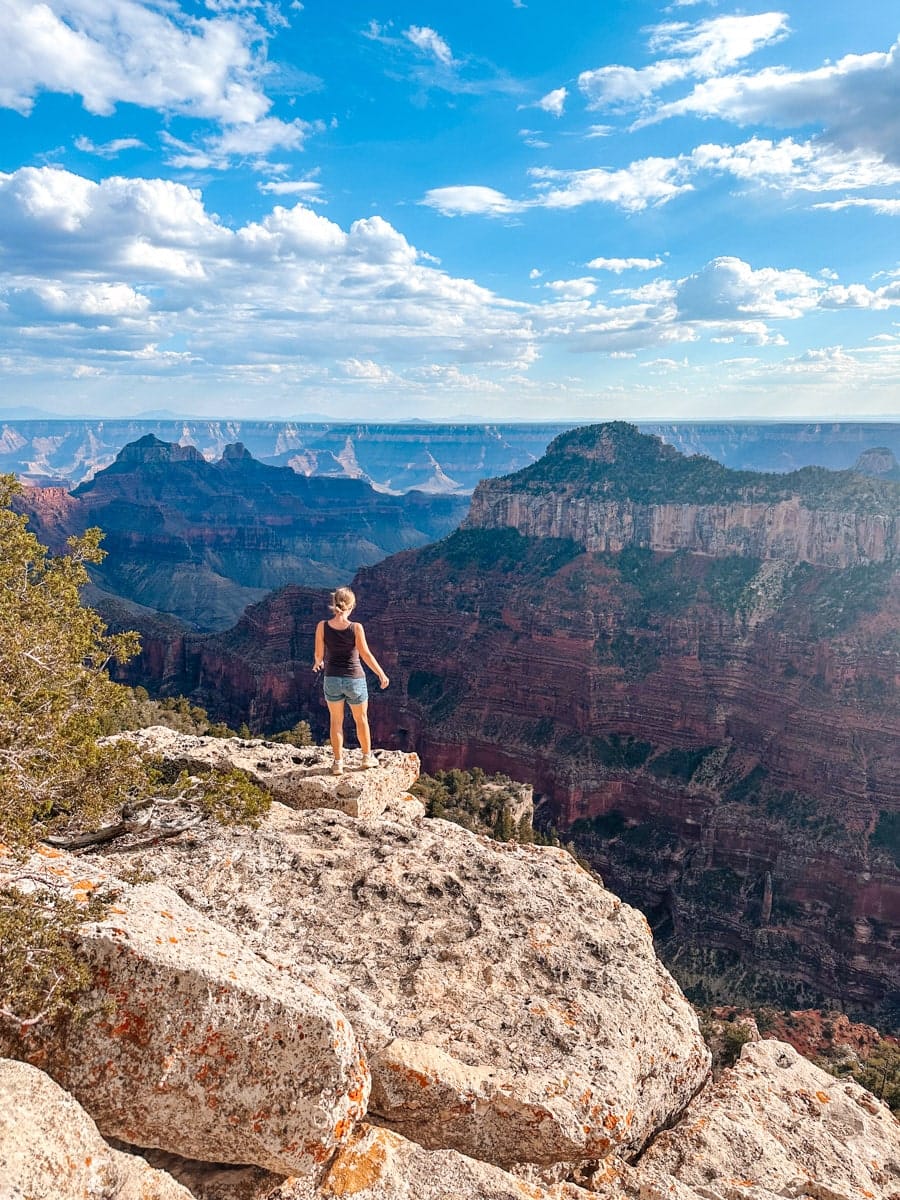 A woman in bright athletic wear hikes along a narrow dirt trail at Bryce Canyon National Park, Utah. The path hugs the edge of a cliff overlooking an unreal landscape of orange hoodoos, spires, and distant mesas under a cloudless sky.