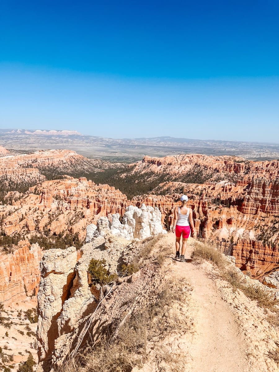 A woman in bright athletic wear hikes along a narrow dirt trail at Bryce Canyon National Park, Utah. The path hugs the edge of a cliff overlooking an unreal landscape of orange hoodoos, spires, and distant mesas under a cloudless sky.