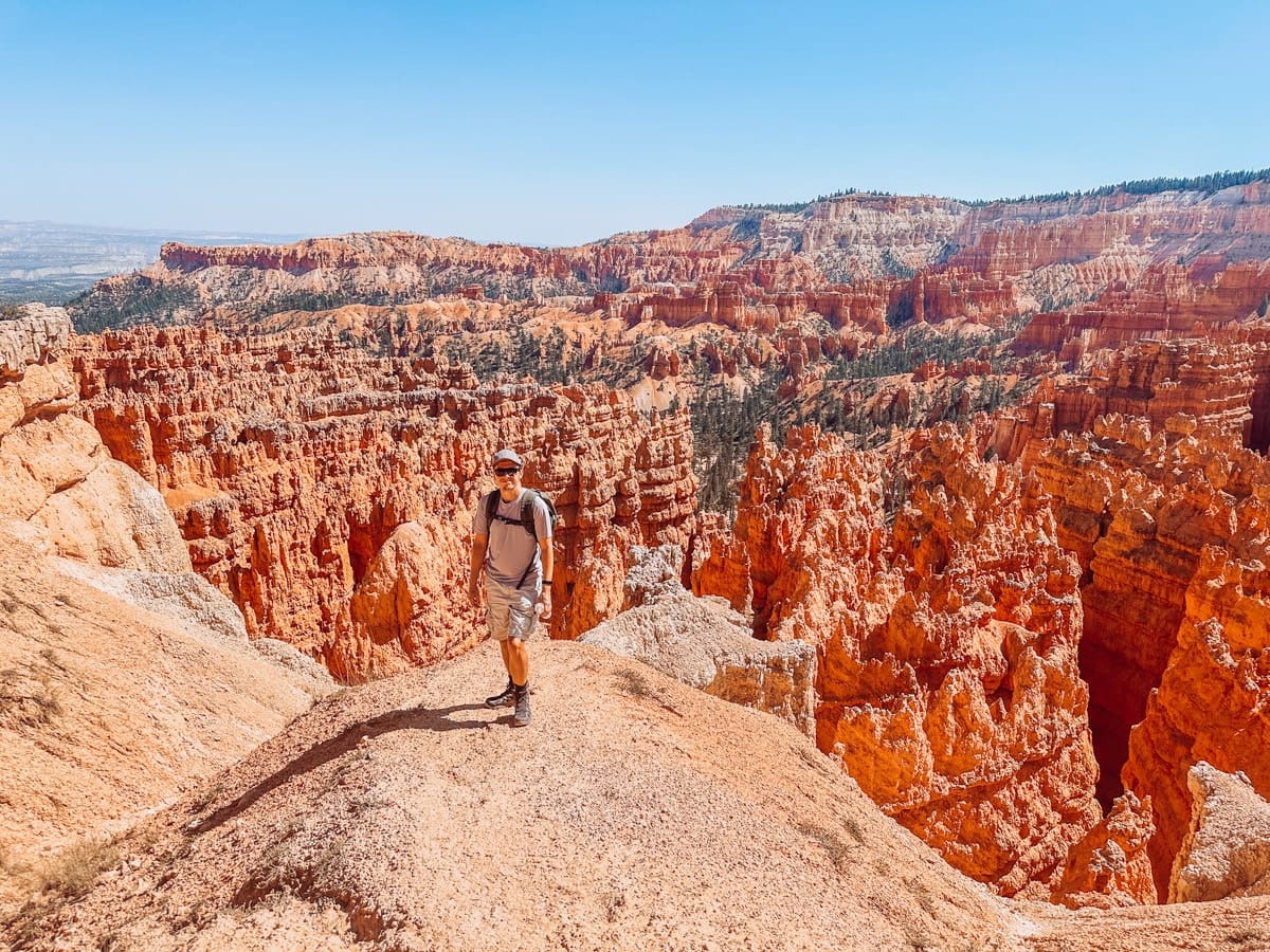 A hiker stands on a trail surrounded by the iconic red rock hoodoos of Bryce Canyon. The towering spires create a maze-like landscape that glows orange in the sunlight, contrasting with the blue sky and green pine trees peeking through the canyon.