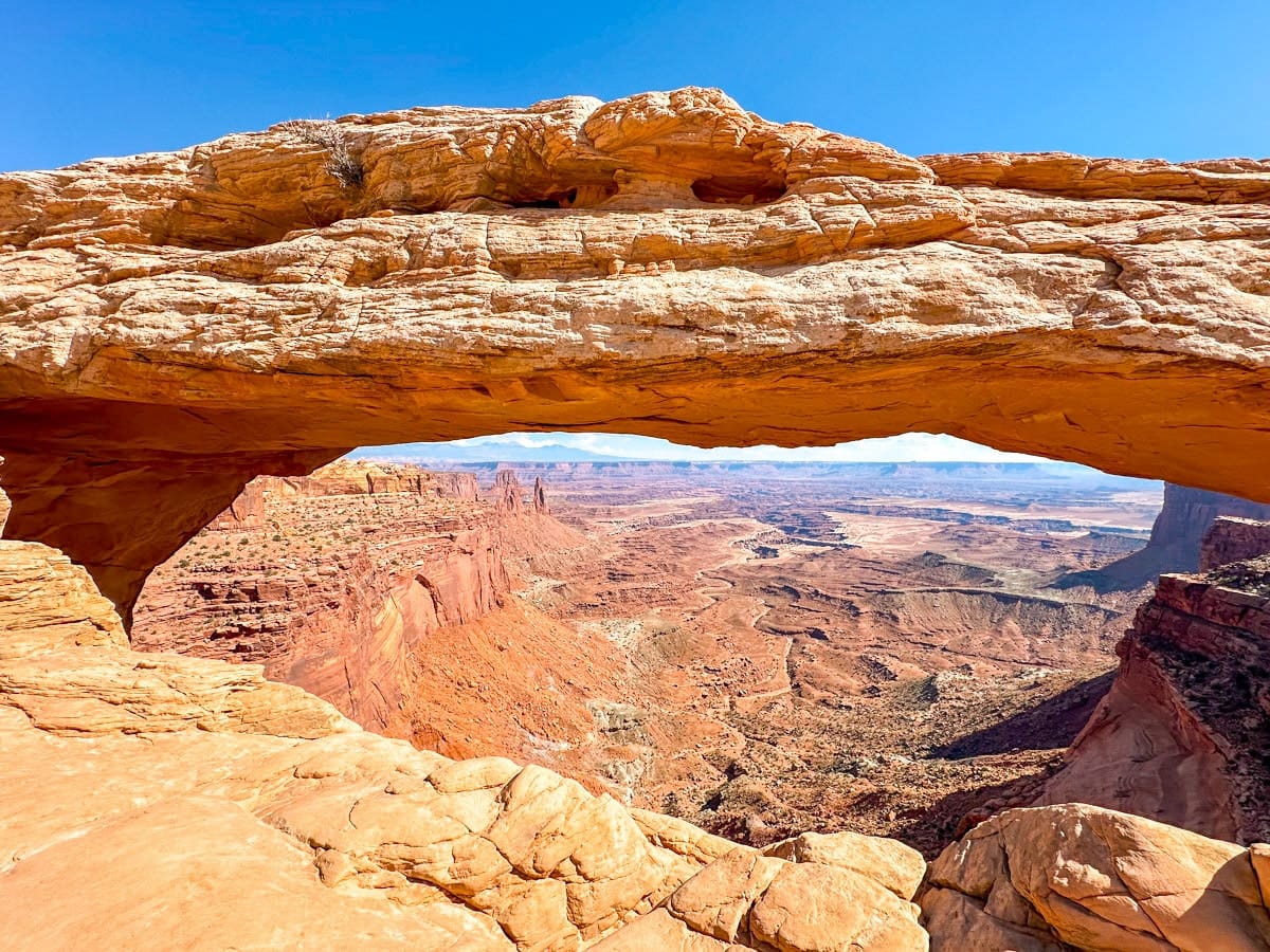 A spectacular view through Mesa Arch in Canyonlands National Park, Utah, looking out over a vast, rugged landscape of canyons, cliffs, and red rock formations. The arch frames the scenery like a natural window, lit beautifully under a vibrant blue sky.
