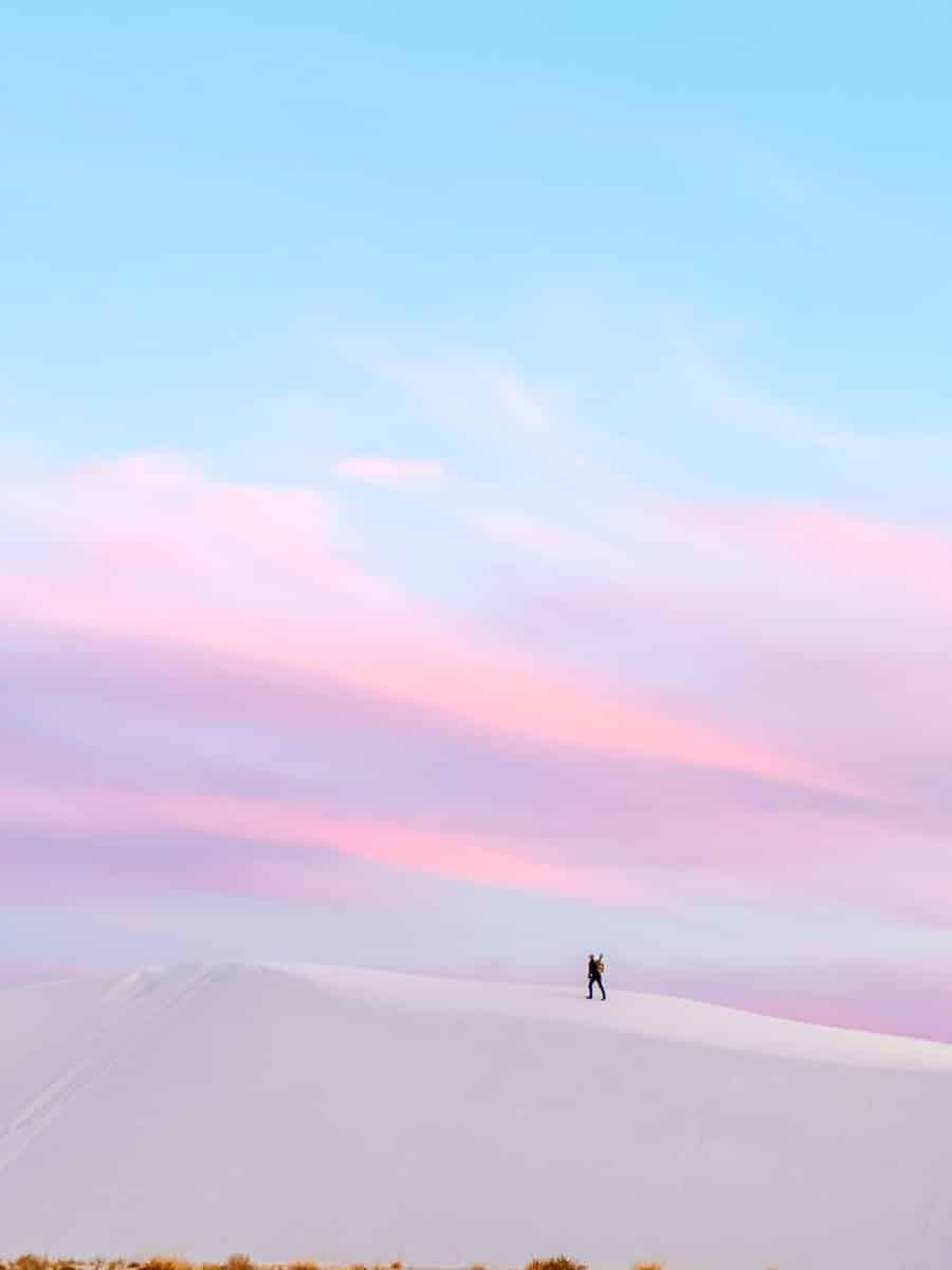 A tiny silhouette of a person stands on top of a massive white dune in White Sands National Park, framed by a pastel sky painted in shades of pink, lavender, and baby blue. The smooth texture of the untouched dune glows under the fading light, creating a peaceful and surreal moment.