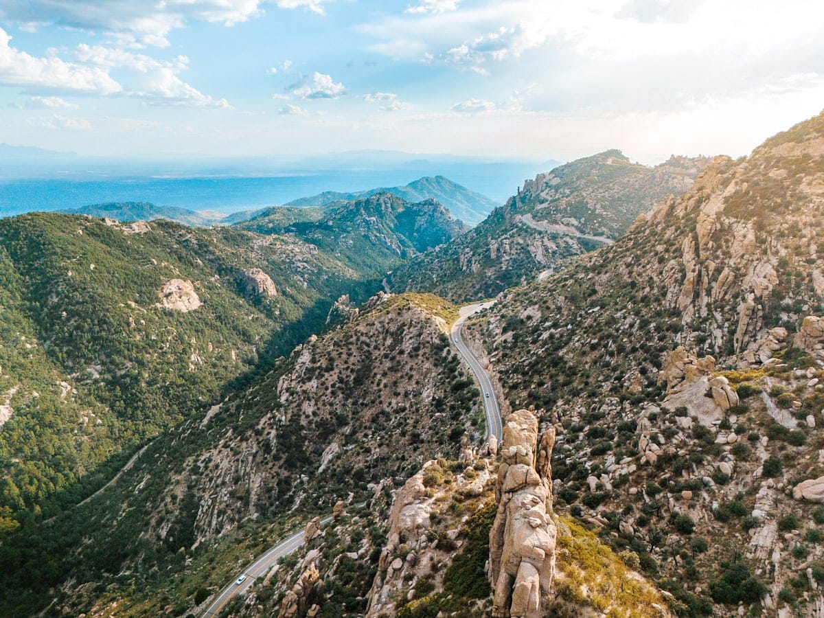 A breathtaking aerial view of the winding road through Mount Lemmon in southern Arizona, with dramatic rock formations, dense green forest, and distant blue haze on the horizon. The golden sunlight highlights the jagged cliffs and curving highway, showcasing one of the most beautiful drives in the Southwest.