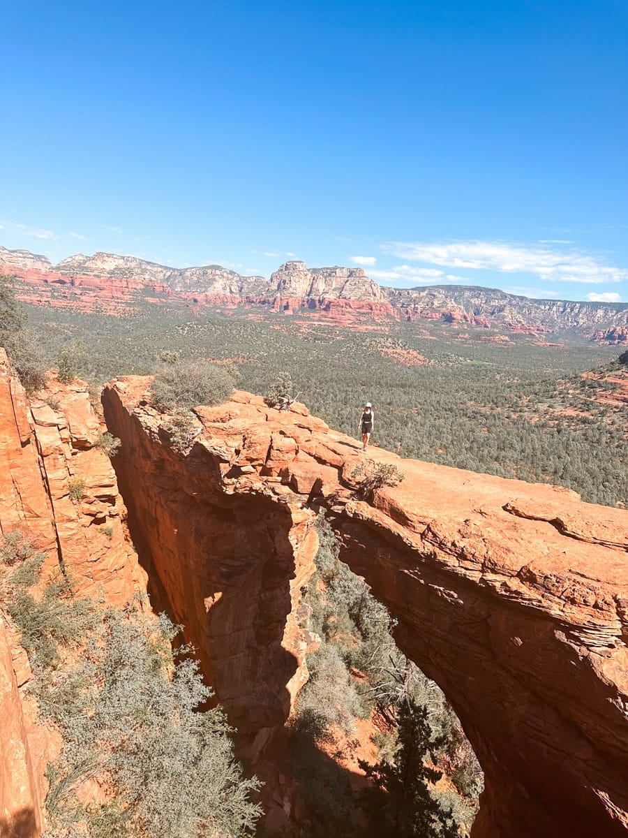 A lone hiker stands on top of Devil’s Bridge, a natural red rock arch surrounded by pine trees and sweeping views of the red rock cliffs in Sedona, Arizona. The sky is a clear, crisp blue, and the rugged terrain below adds depth and drama to this adventurous scene.