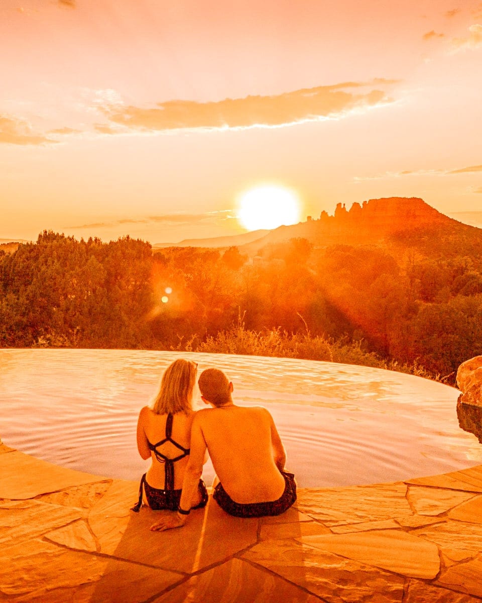 A romantic couple sits side by side on the edge of a cliffside infinity pool, watching the sun set behind Sedona’s red rock formations. The sky and water are drenched in rich orange hues, while the couple’s silhouettes are softly backlit, creating a warm and intimate desert moment.