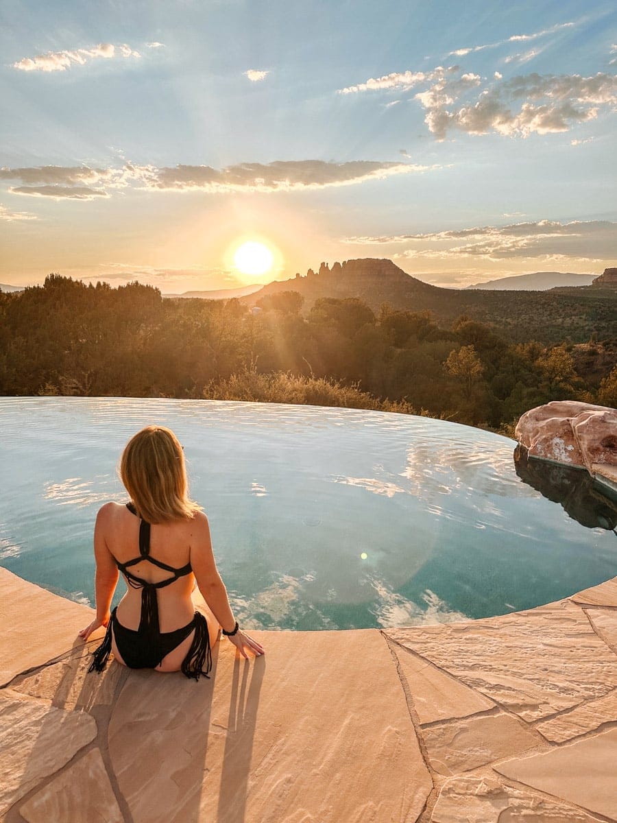 A woman in a black strappy swimsuit sits at the edge of a serene infinity pool in Sedona, gazing out at the glowing red rock formations during sunset. The sky is scattered with golden clouds, and the reflection on the water mirrors the tranquil beauty of the surrounding landscape.