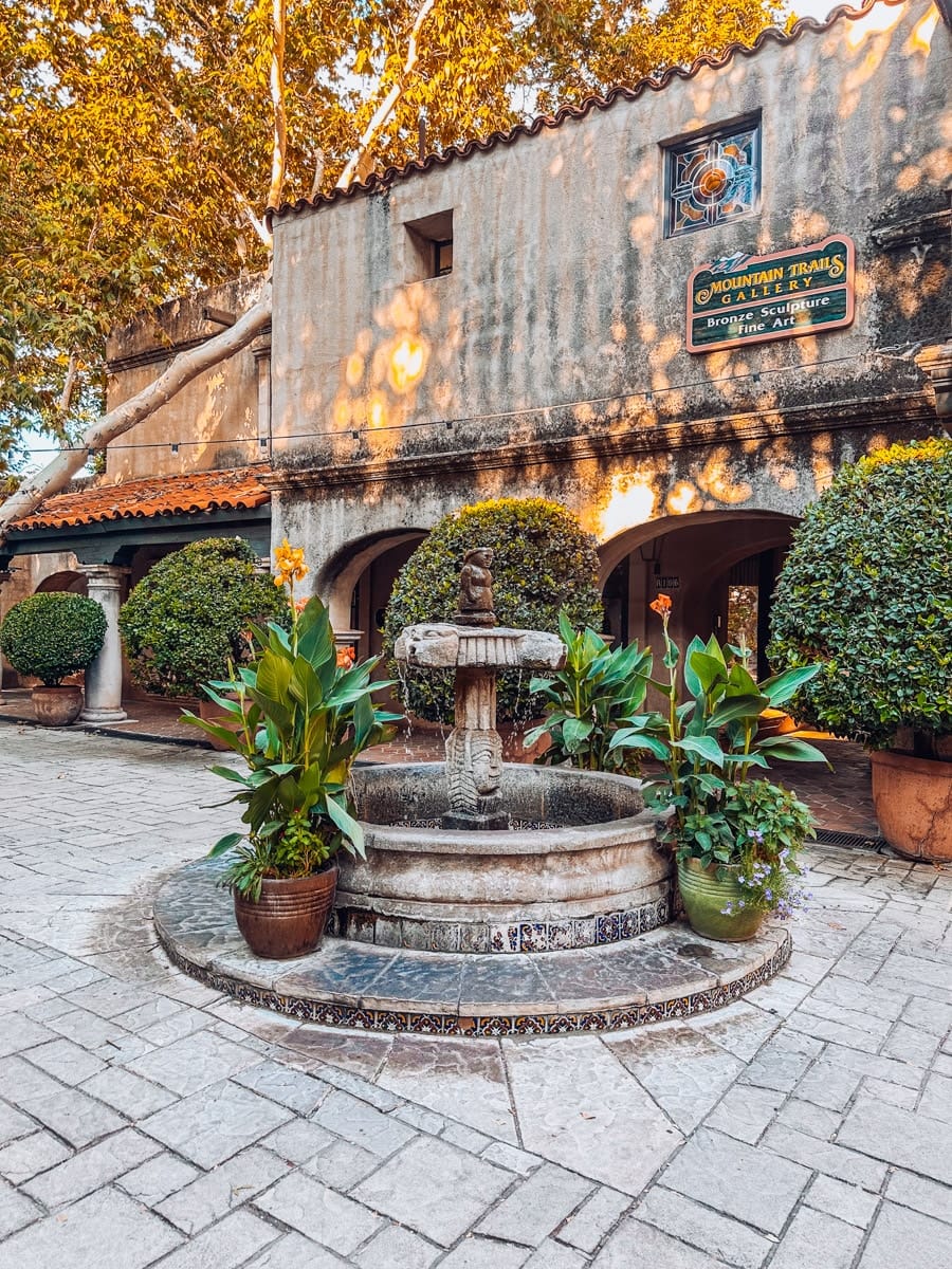 A charming courtyard scene outside Mountain Trails Gallery in Sedona, Arizona, featuring a classic stone fountain surrounded by lush potted plants and Spanish-style architecture. The building’s tan stucco walls are kissed with warm fall light, and yellow leaves peek through the trees above, adding cozy seasonal vibes.
