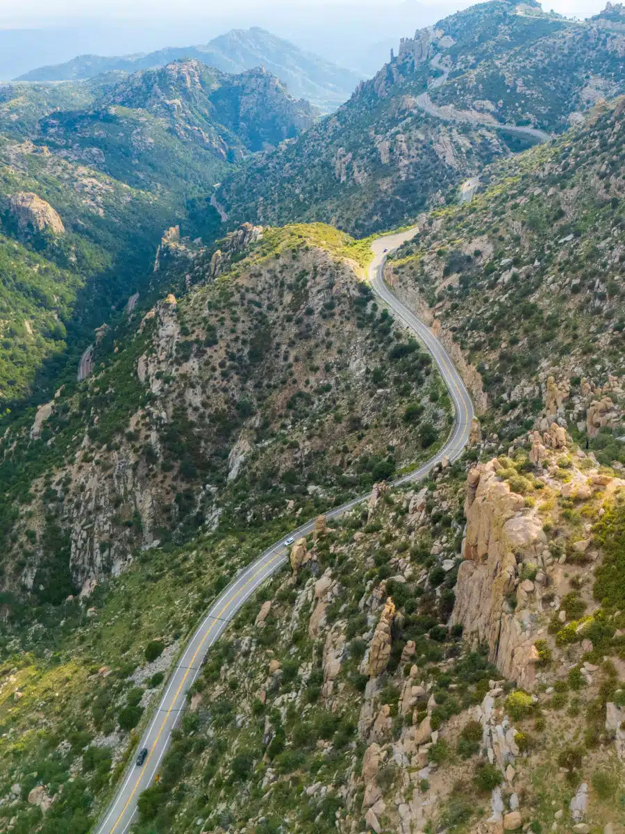 Stunning mountain road cutting through rocky peaks and forested slopes of Mount Lemmon in Arizona, with a view stretching toward the hazy desert valley below.