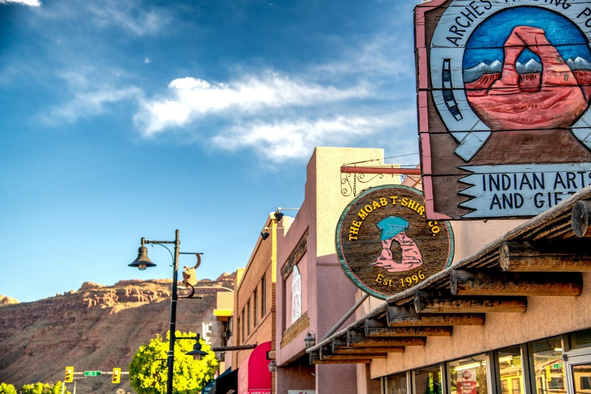 Colorful storefront signs in downtown Moab, Utah, including a round “Moab T-Shirt Co.” sign and an “Indian Arts and Gifts” mural, with red rock cliffs in the background under a bright blue sky.