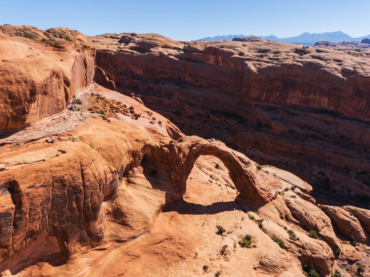 Aerial view of Corona Arch nestled in the red rocks near Moab, Utah, surrounded by desert terrain and steep sandstone cliffs.