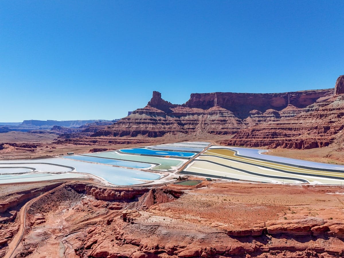 Aerial view of the colorful potash evaporation ponds near Moab, Utah, set against a dramatic red rock canyon and a towering mesa under a clear blue sky.