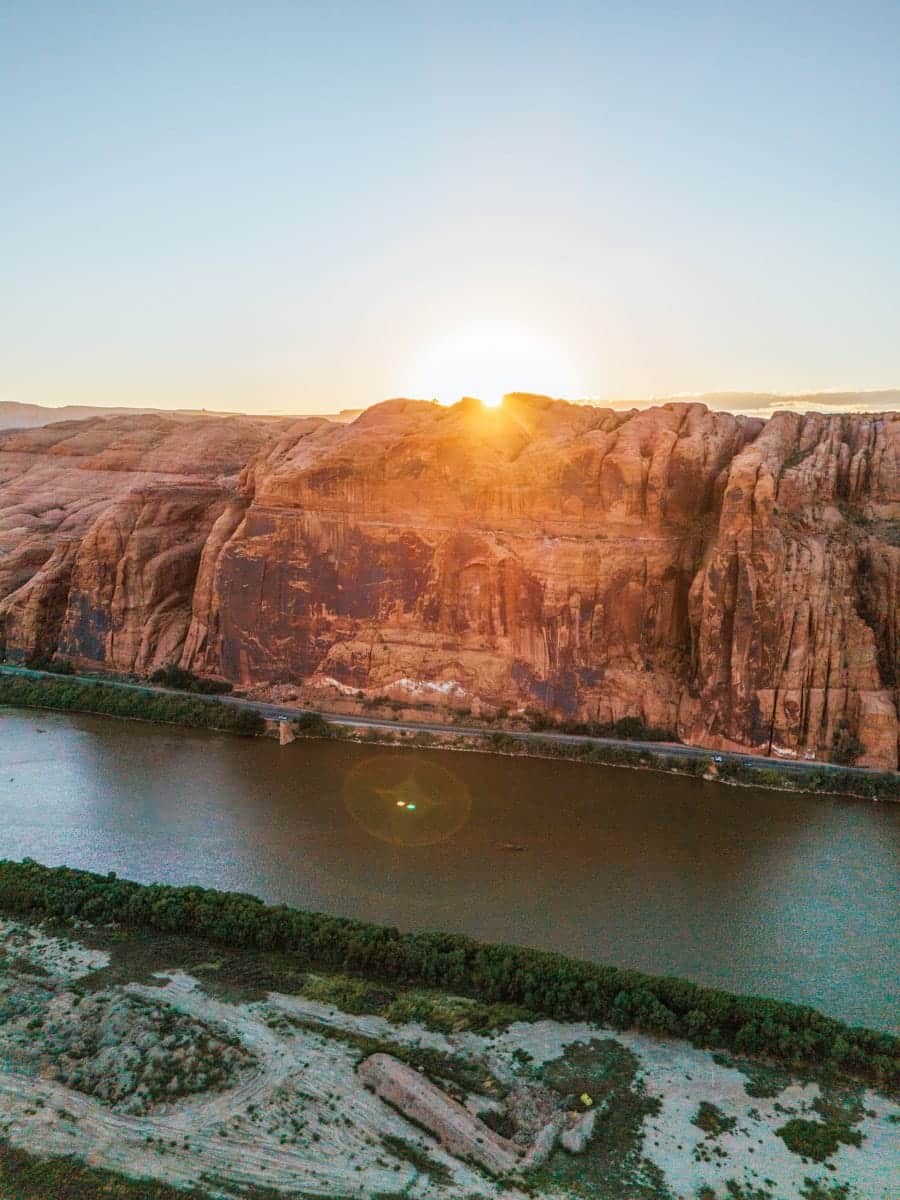 Sunrise peeking over red sandstone cliffs in Moab, Utah, casting golden light on the Colorado River below in a serene desert landscape.