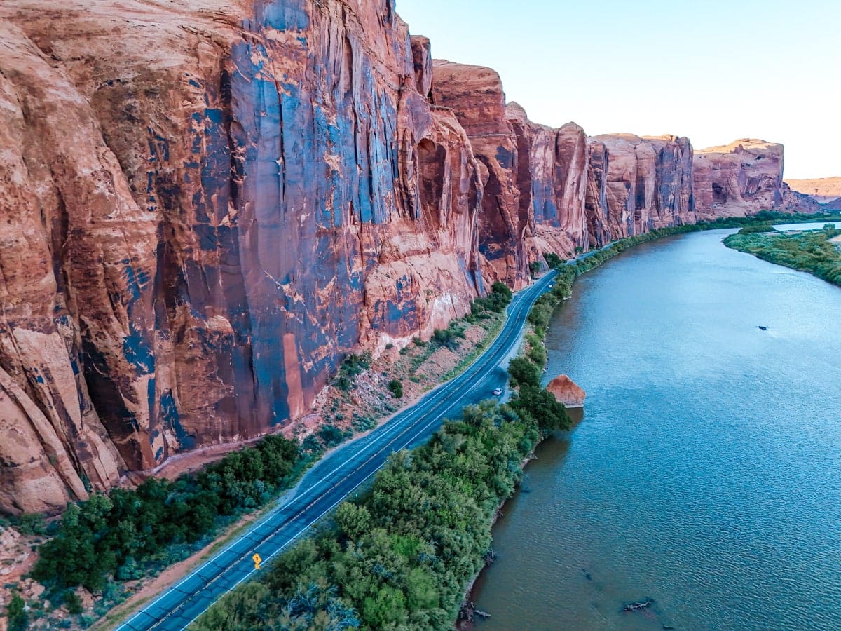Aerial view of the scenic Potash Road hugging the Colorado River, with massive sandstone cliffs towering on one side and lush greenery lining the riverbank.
