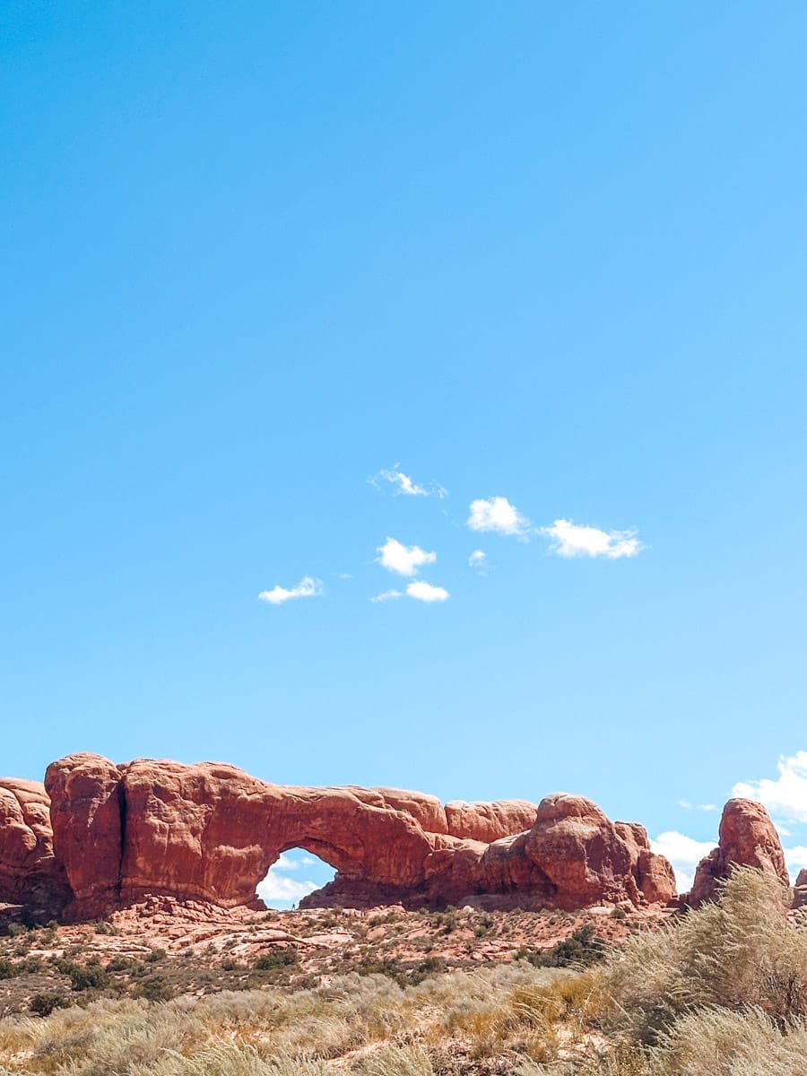 Wide view of the North Window Arch in Arches National Park, Utah, framed by desert brush and a bright blue sky with a few puffy clouds.