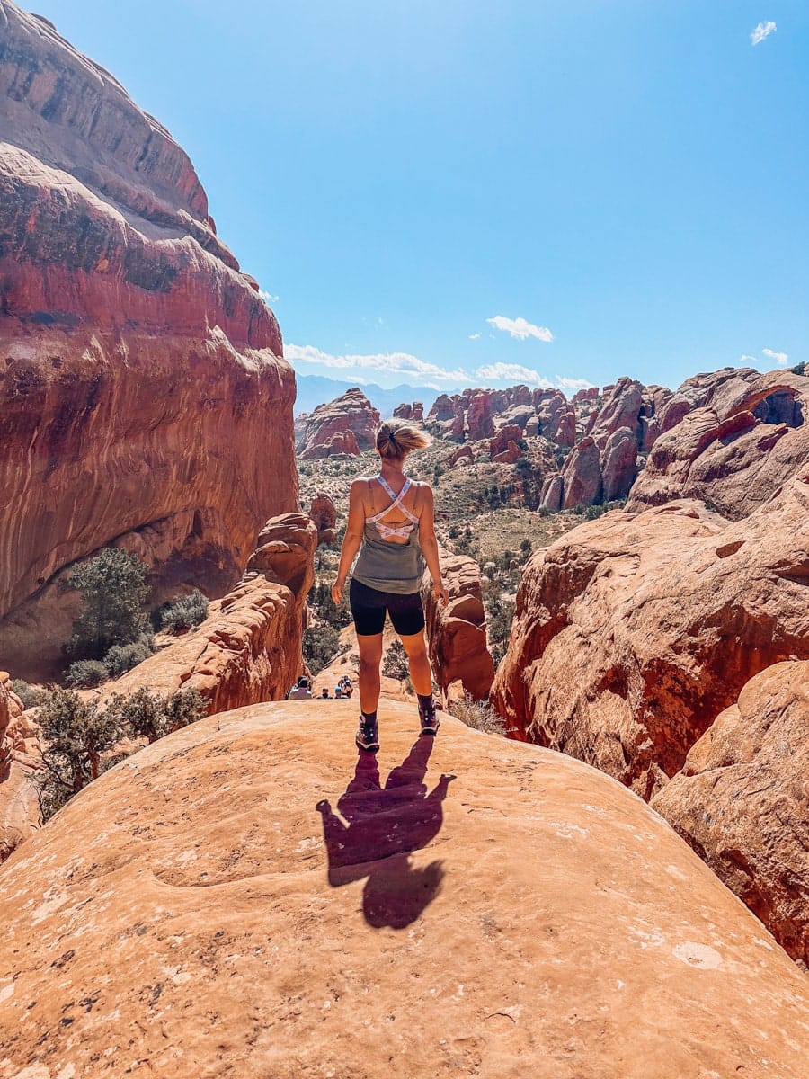 Back view of a woman standing confidently on top of a red sandstone dome, surrounded by rugged canyon walls in Arches National Park.