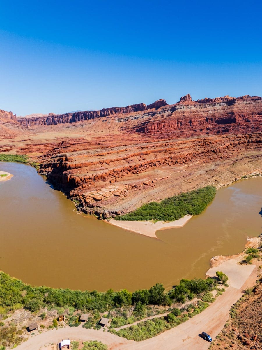 Aerial view of a dramatic bend in the Colorado River surrounded by striped red rock formations and dirt roads near Moab, Utah.