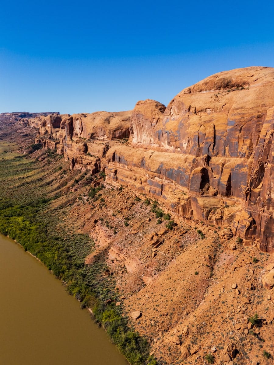 Drone photo of the Colorado River running beside towering red cliffs and desert vegetation near Moab, Utah, on a sunny day.