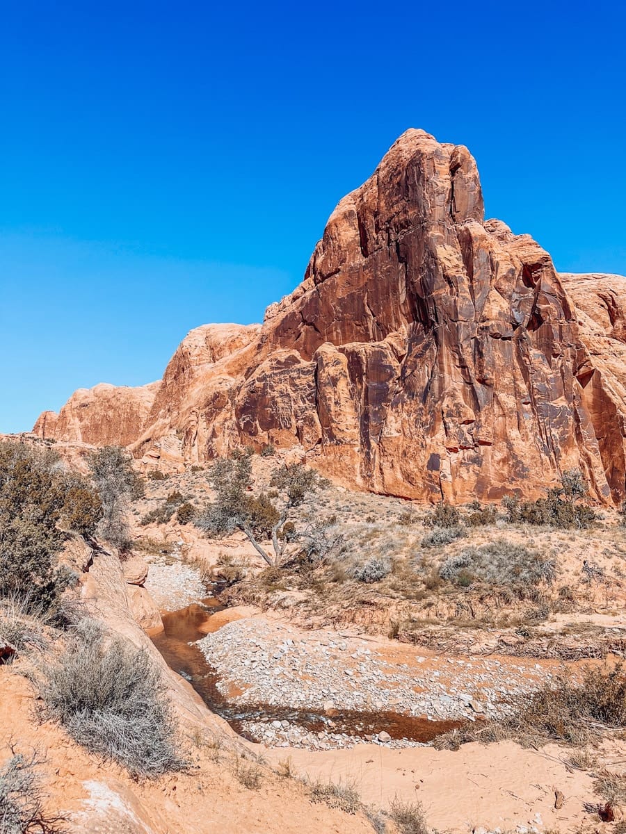 A scenic desert landscape in Utah with a sharp sandstone peak rising beside a small stream and scattered desert shrubs under a vibrant blue sky.