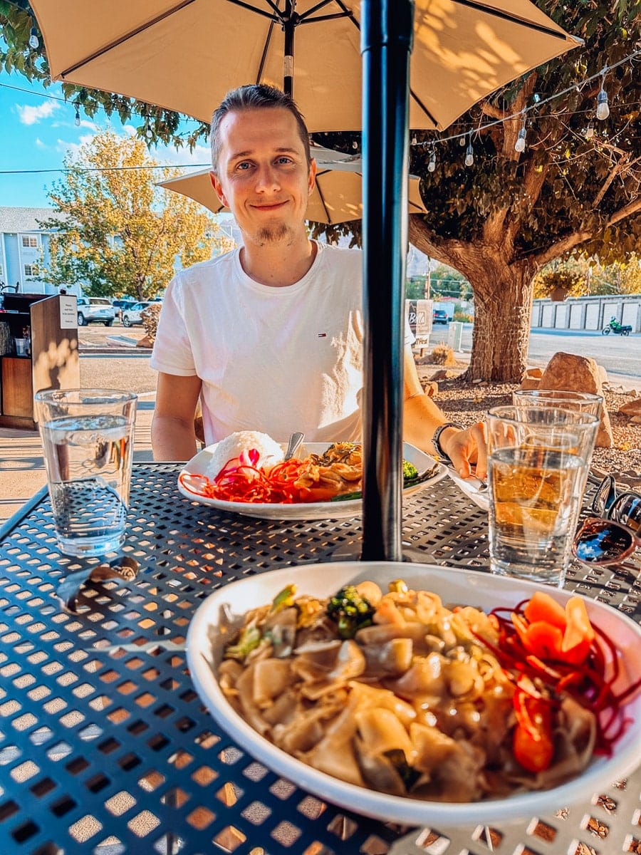 Smiling man enjoying a colorful Thai lunch at an outdoor café in Moab, Utah, with umbrellas, string lights, and fall trees in the background.