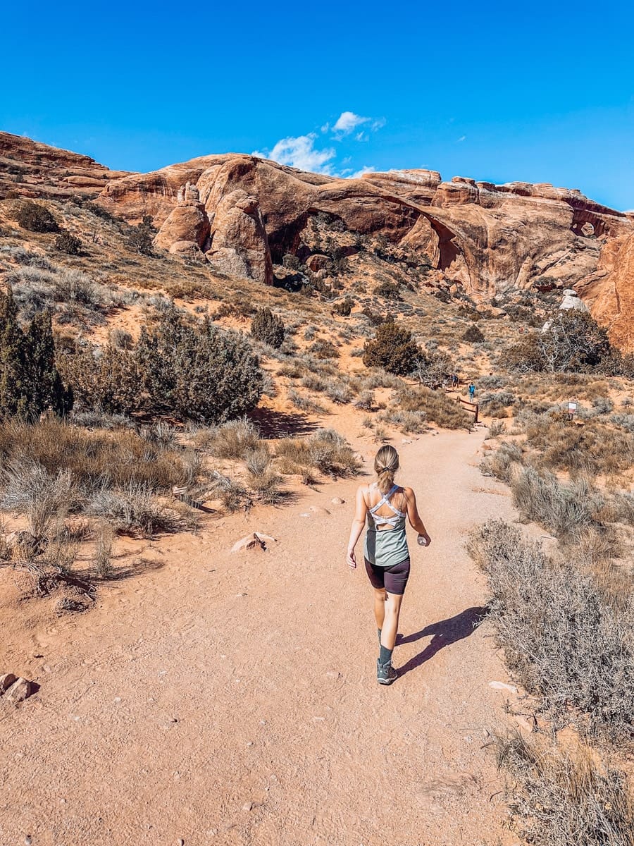 Back view of a hiker walking toward Landscape Arch in Arches National Park, with red rock walls and desert plants along the trail.