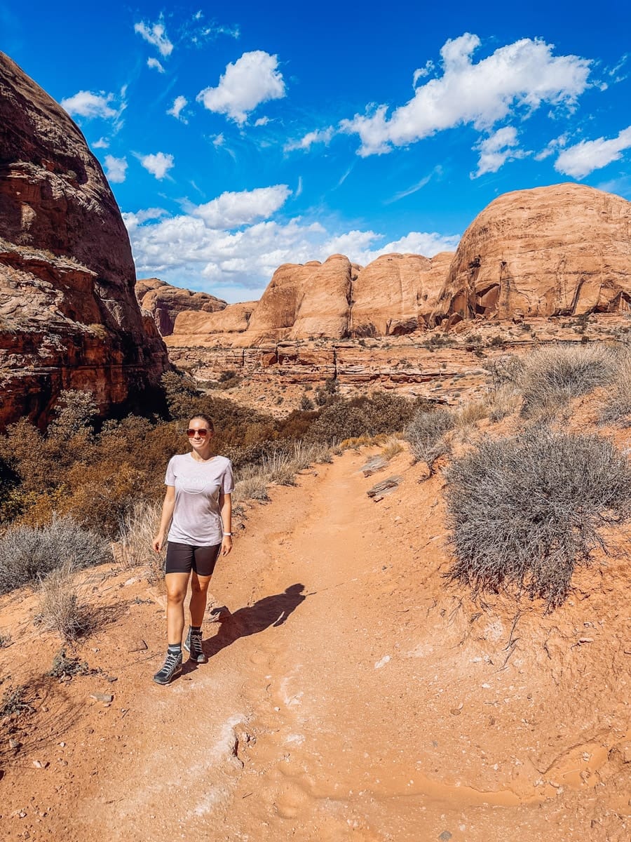 Woman hiking a scenic desert trail surrounded by tall red rock formations under a bright blue sky in Moab, Utah.