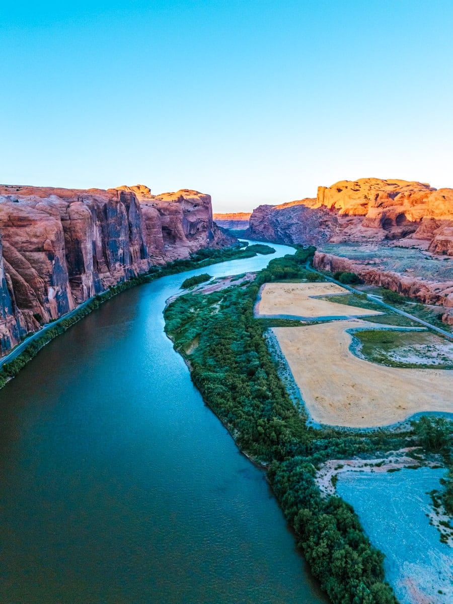 Aerial view of the Colorado River curving between towering red rock cliffs and green farmland near Moab, Utah, at golden hour.