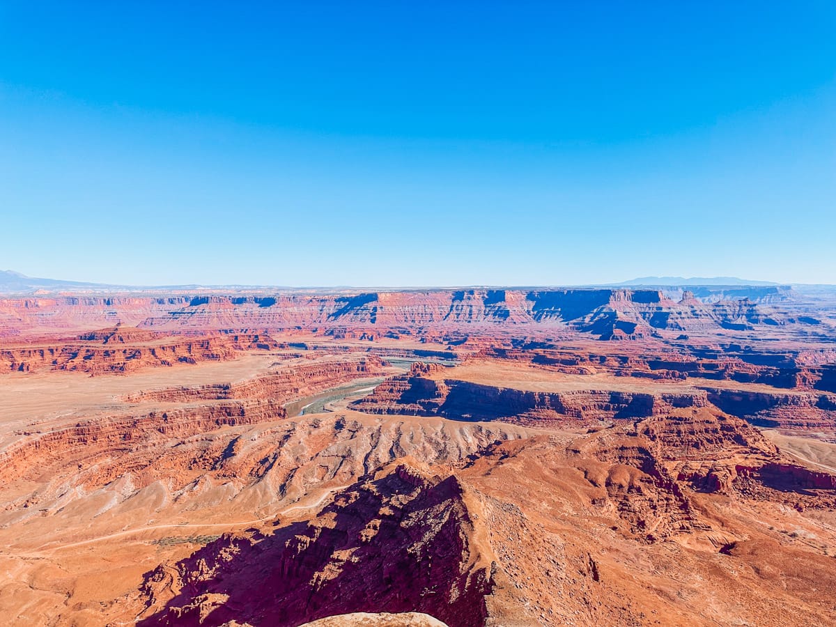 Stunning panoramic view of Dead Horse Point State Park with layered red rock canyons and the winding Colorado River far below, captured under a bright blue sky.