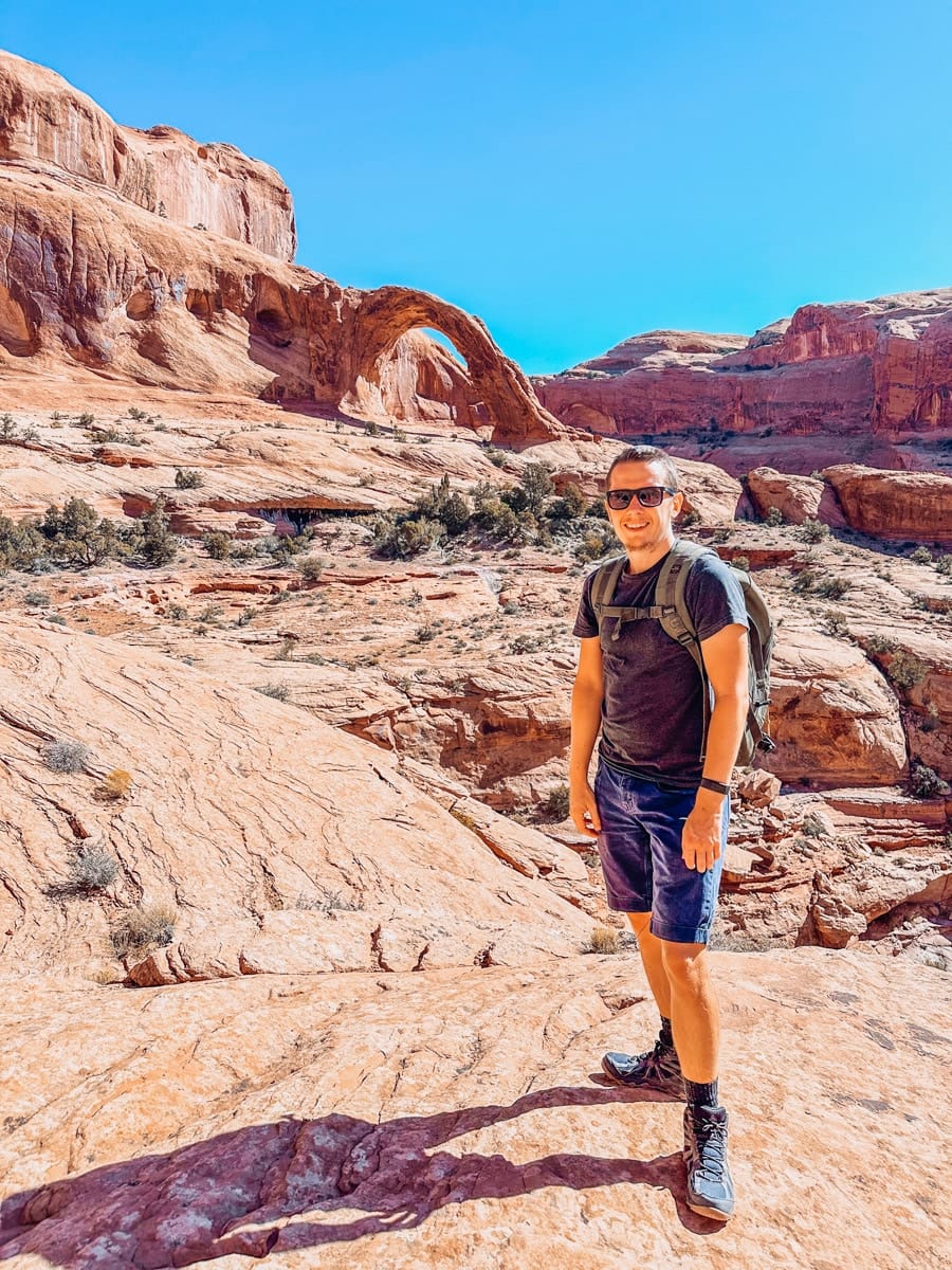 The author, a smiling man in hiking gear and sunglasses, stands on red slickrock with Corona Arch in the background during a sunny day of adventure in Moab.