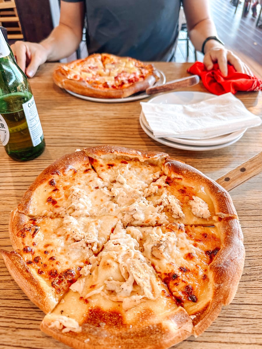 Freshly baked cheese and chicken pizza served on a wooden table, with a second pizza and a cold beer in front of the author in the background.