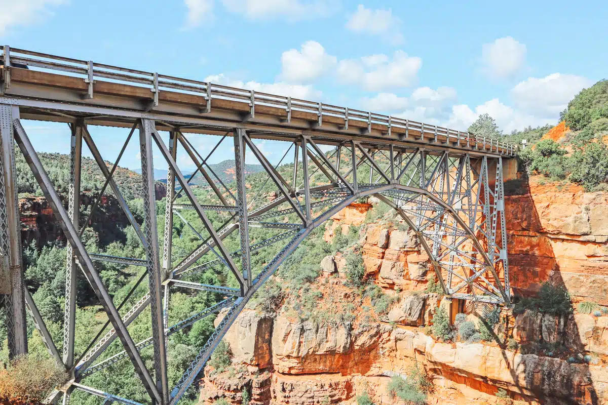 Historic steel bridge stretching across Oak Creek Canyon in Arizona, framed by red rock cliffs and lush green forest under a bright blue sky.