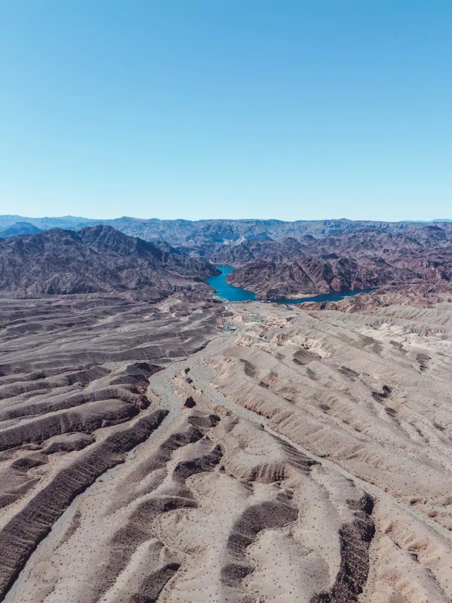  Drone shot of winding ridges in the Nevada desert leading to the blue waters of Lake Mead, surrounded by jagged mountains and a clear blue sky.