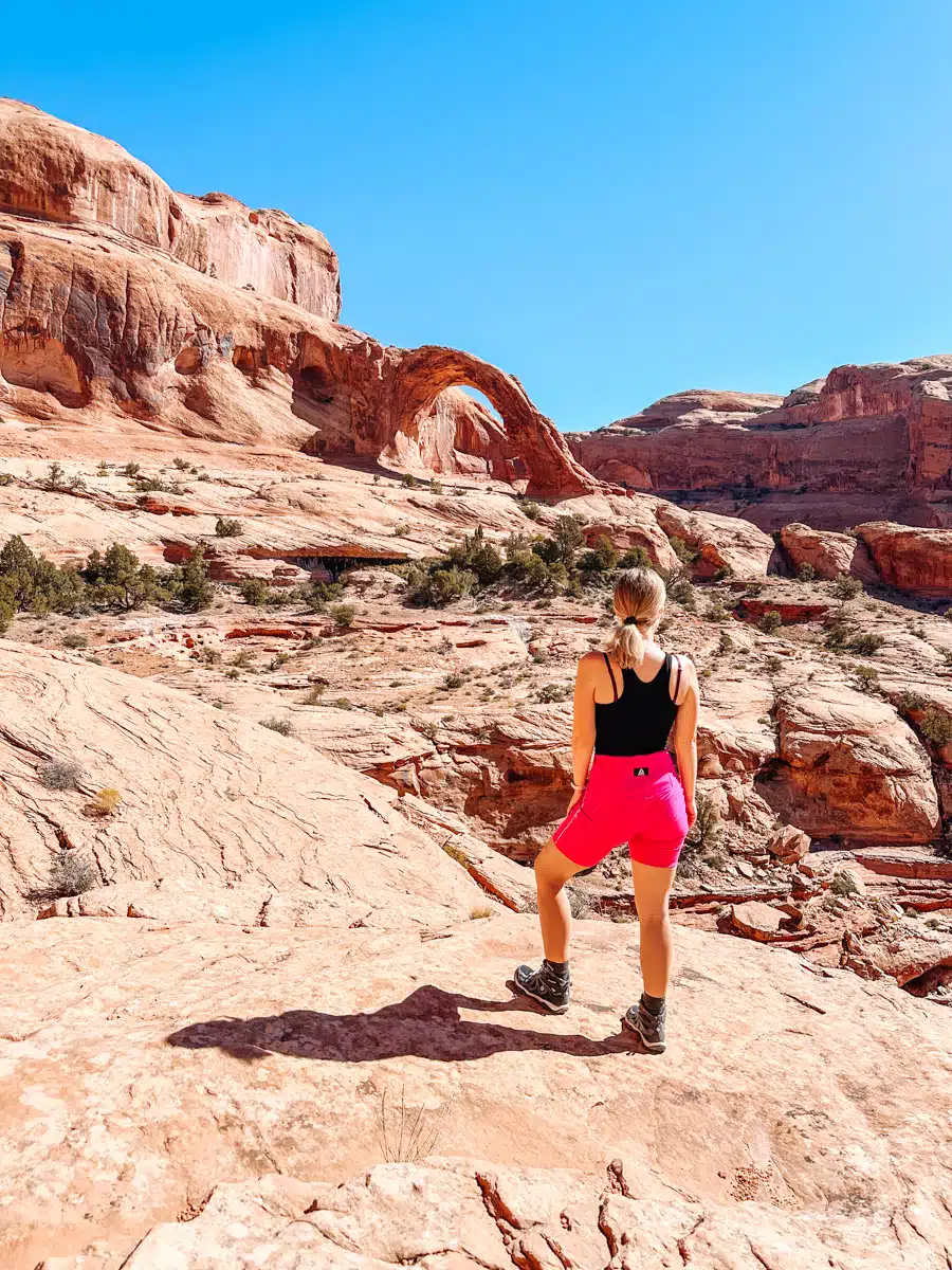 Woman in pink shorts standing on slickrock, gazing at Corona Arch under a bright blue sky in Moab, Utah.