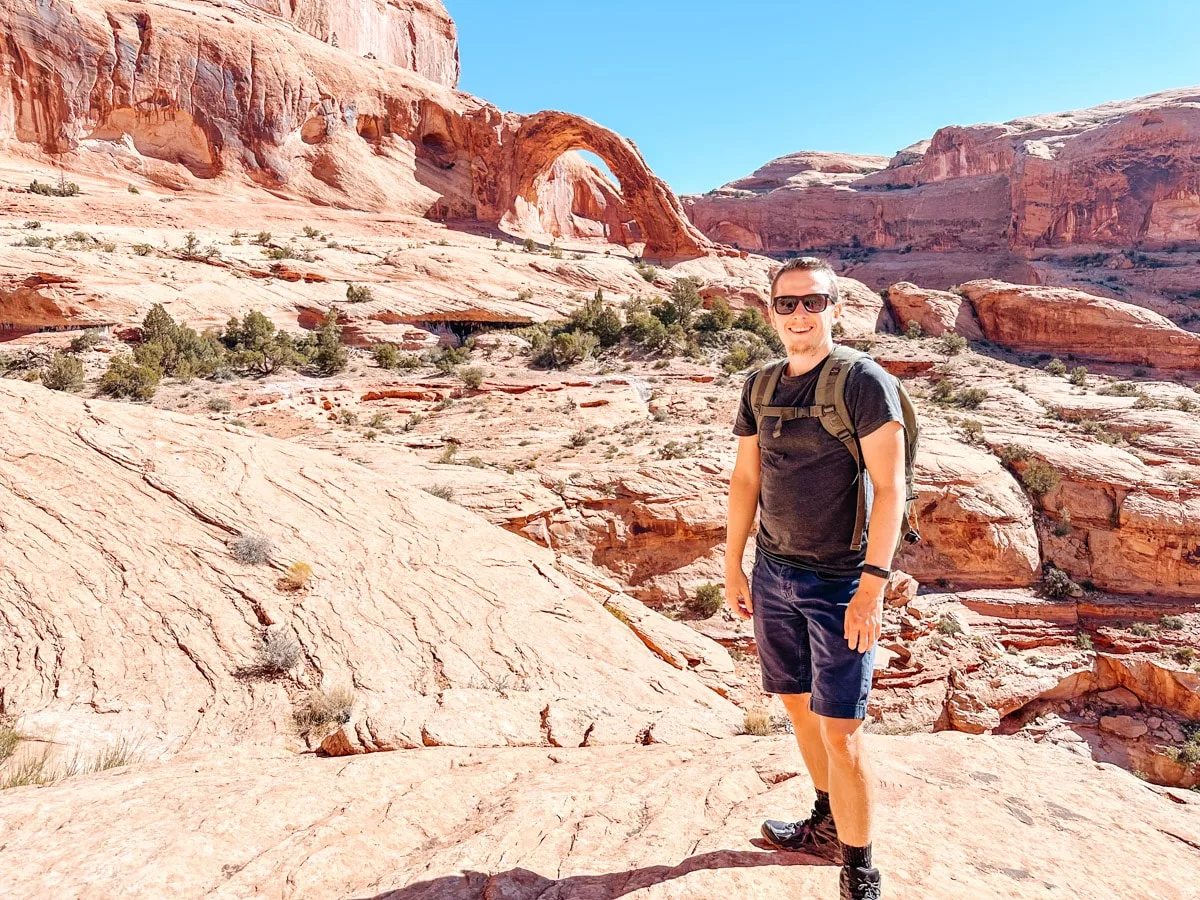 Man in hiking gear smiling in front of the red rock landscape and Corona Arch in the distance, Moab, Utah.