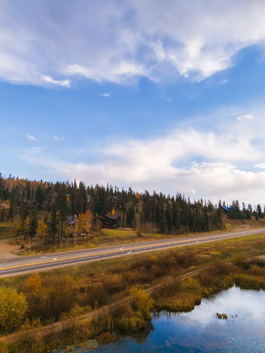 Peaceful roadside scene with a pond and forested hills glowing with early autumn colors under a soft sky.