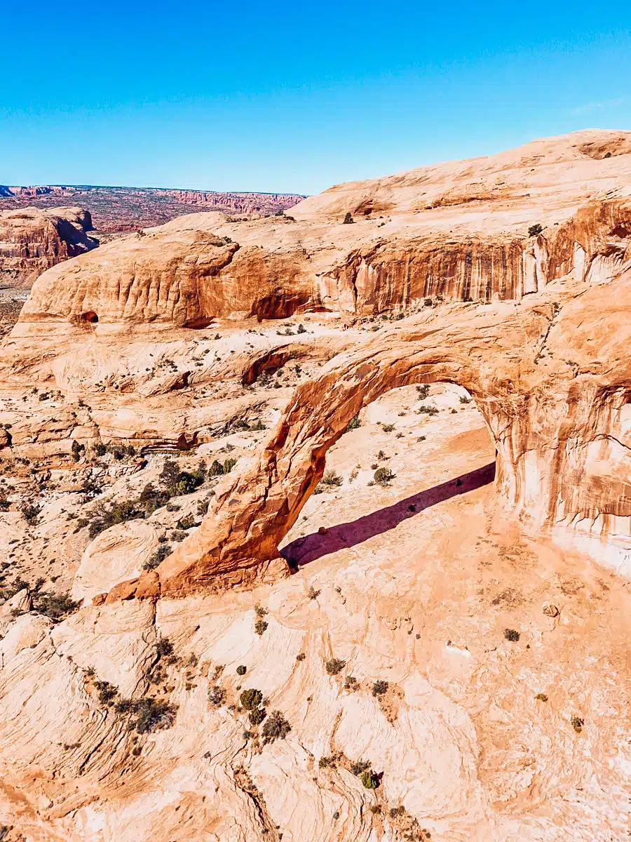 Drone view of Corona Arch curving over desert sandstone, casting a long shadow on the slickrock floor below.