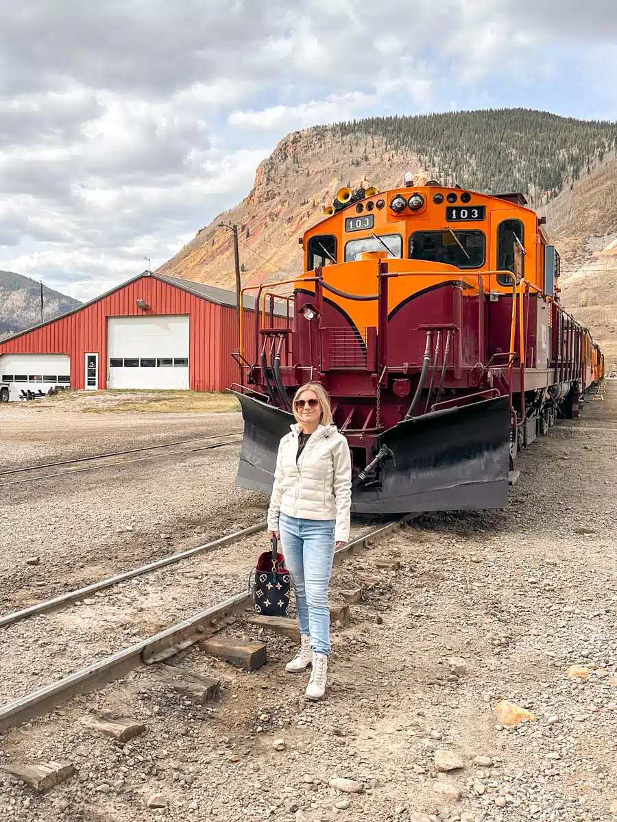 Woman in a white jacket posing in front of the orange and maroon train engine in Silverton, Colorado, ready for an adventure.